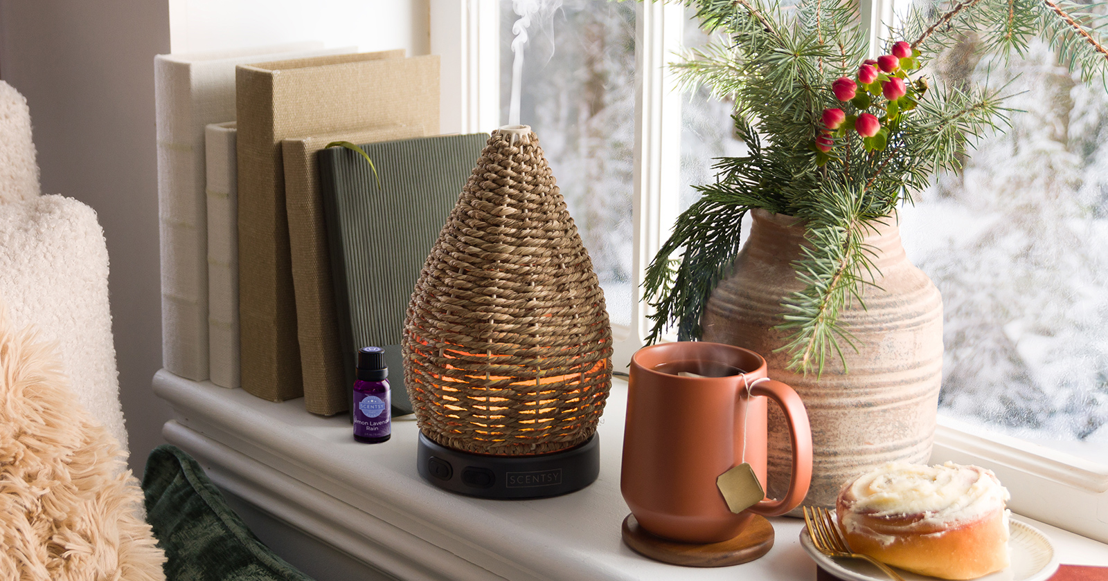 Scentsy Wonder woven rattan teardrop essential oil diffuser with warm amber glow on white windowsill, displayed with terracotta mug, pine branch arrangement, books, and frosted cinnamon roll