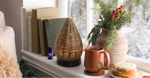 Scentsy Wonder woven rattan teardrop essential oil diffuser with warm amber glow on white windowsill, displayed with terracotta mug, pine branch arrangement, books, and frosted cinnamon roll