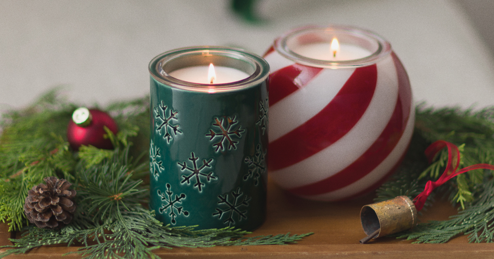A green candle holder etched with snowflakes and a red-and-white striped candy cane swirl candle holder, both lit, styled with pine garland and holiday ornaments for October holiday merchandising.