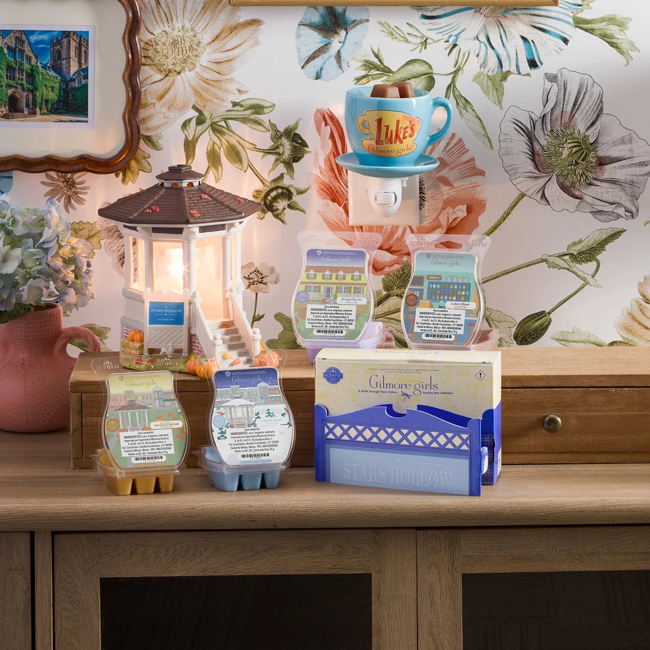 El Calentador Scentsy – The Gilmore Girls: Welcome to Stars Hollow y las Barras Scentsy de la Colección de Cera Scentsy – A Stroll Through Stars Hollow arreglados en una mesa. Detrás de eso, un Mini Calentador Scentsy – Luke’s Coffee Mug está enchufado en una pared con papel pintado floral.