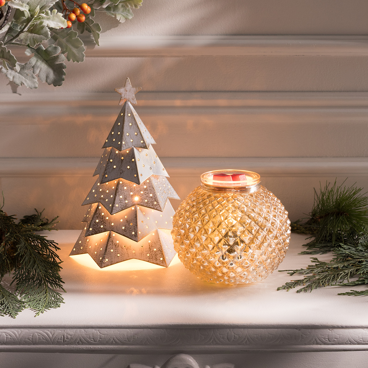 Exhibición festiva con dos calentadores Scentsy de la Colección de Fiestas, que brillan suavemente sobre una repisa decorada con follaje para crear un ambiente acogedor y de temporada.