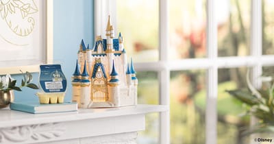 Un Calentador Scentsy – Walt Disney World: Cinderella Castle se muestra junto a una Barra Scentsy – Walt Disney World: Where Dreams Begin en una repisa de chimenea cerca de una ventana.