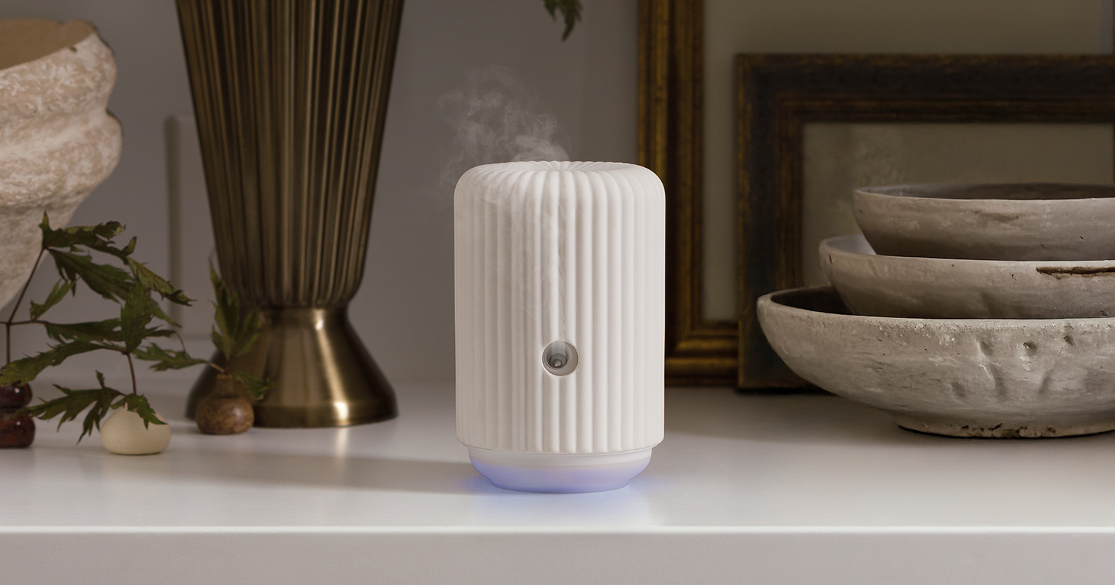  Smart Diffuser in white with a ribbed ceramic design, softly glowing at the base with visible mist rising, styled on a neutral tabletop beside decorative bowls and brass accents.