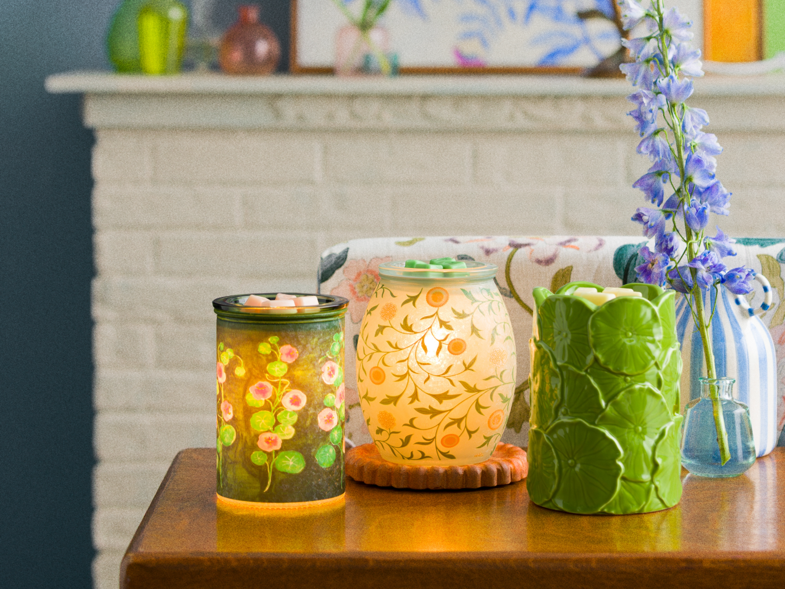 Three decorative Scentsy warmers displayed on a wooden table with wax melts, featuring floral and botanical designs alongside a small vase of flowers.