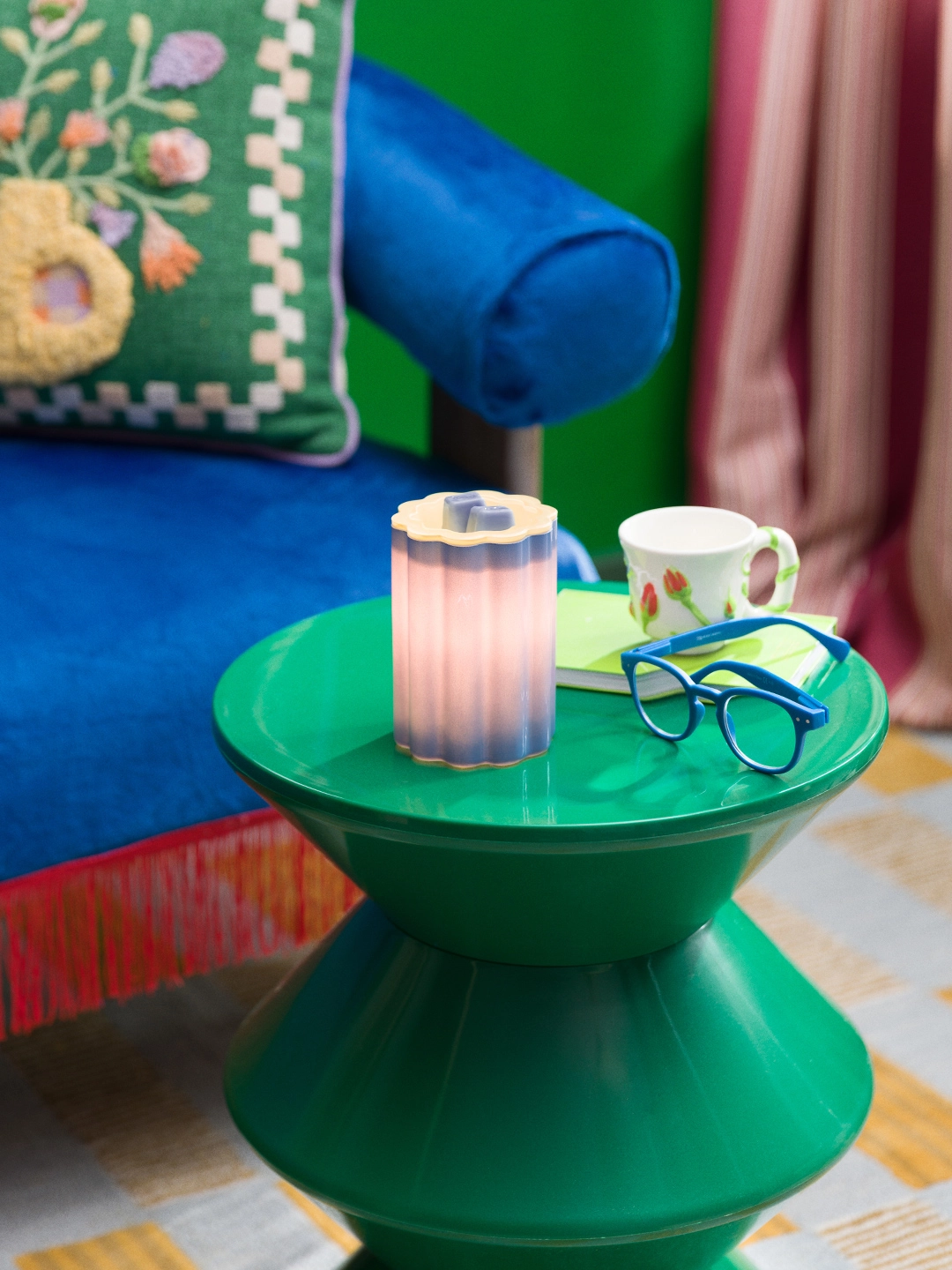 Bloomie-Blue glass warmer lit to reveal a soft blue-to-pink ombré glow, styled on a green side table beside a mug, notebook, and blue glasses.