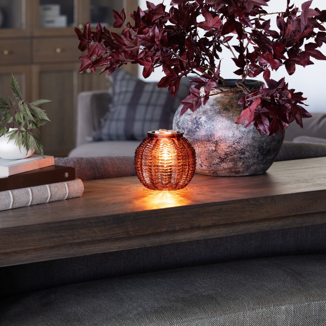 Scentsy Amber Waves Warmer on a table, glowing from within, sitting next to a stack of books and potted plants,