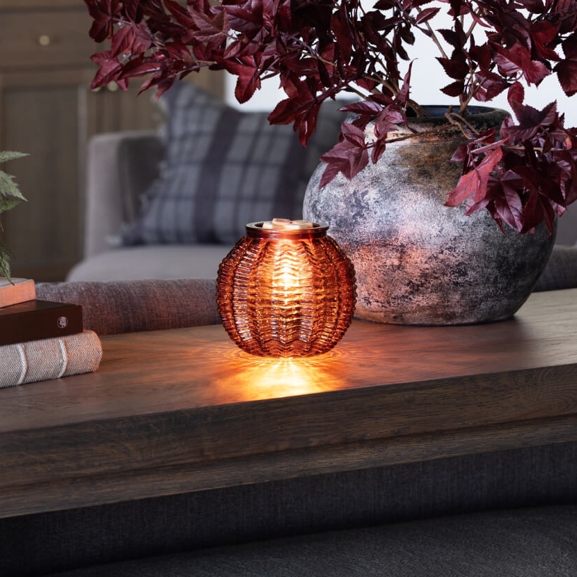 Scentsy Amber Waves Warmer on a table, glowing from within, sitting next to a stack of books and potted plants,