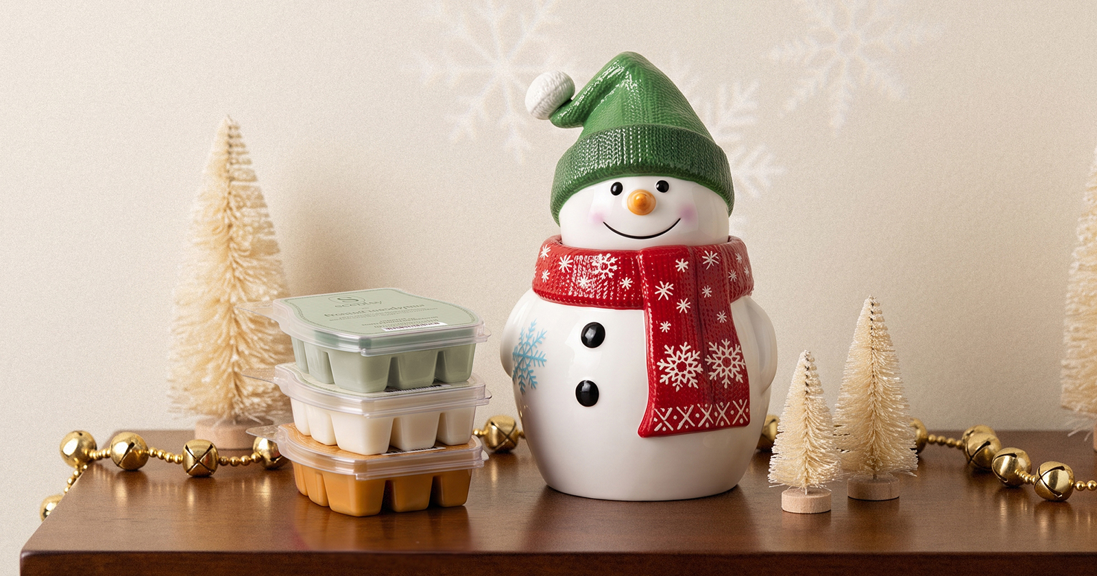 A snowman-shaped Scentsy Warmer wearing a green knit hat and red snowflake scarf is displayed with a stack of Scentsy Bars, bottlebrush trees, and gold jingle bells.