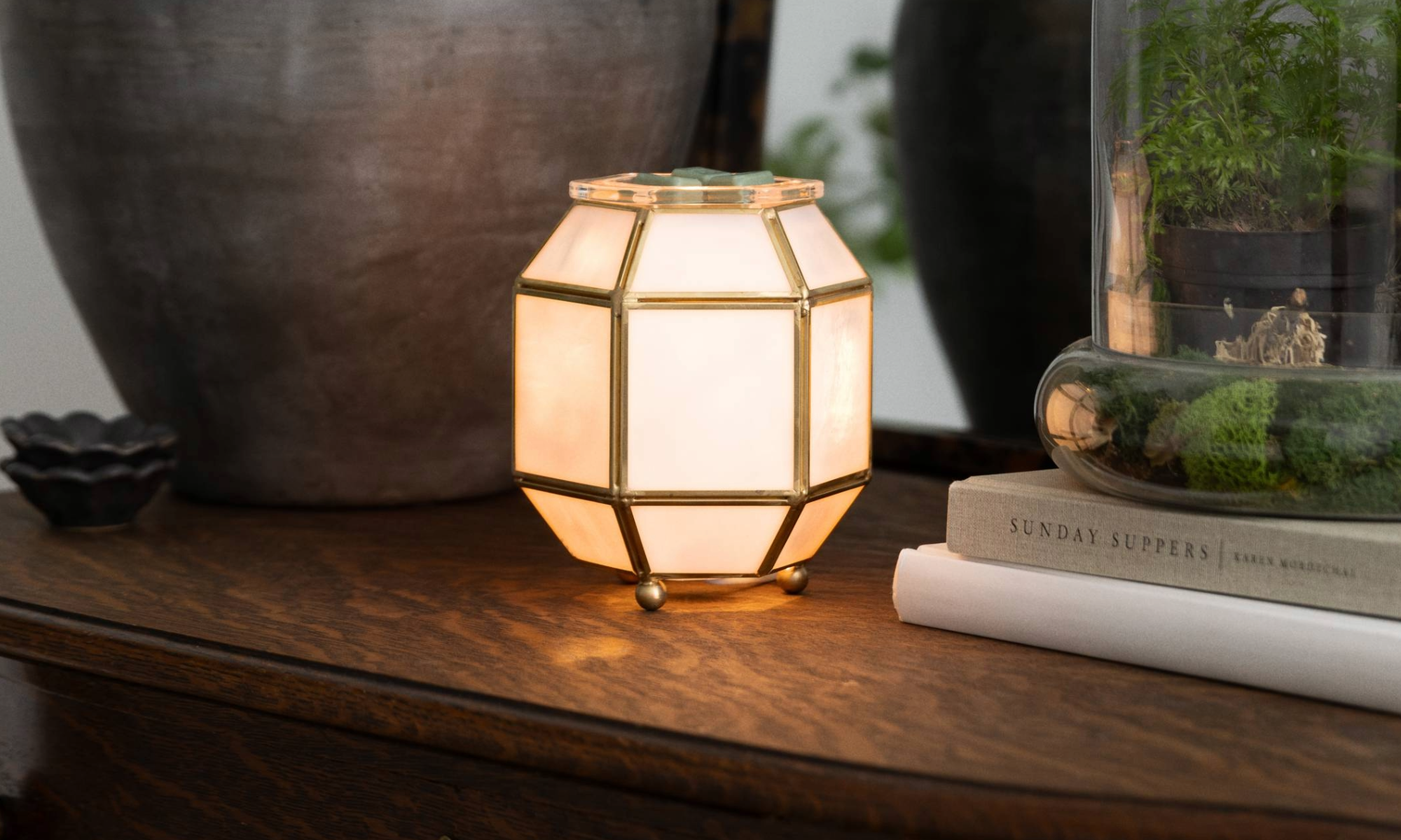 Calentador Clara con paneles geométricos de vidrio y acabado metálico, iluminado sobre una consola de madera junto a plantas verdes y acentos decorativos en un ambiente hogareño cálido y elegante.