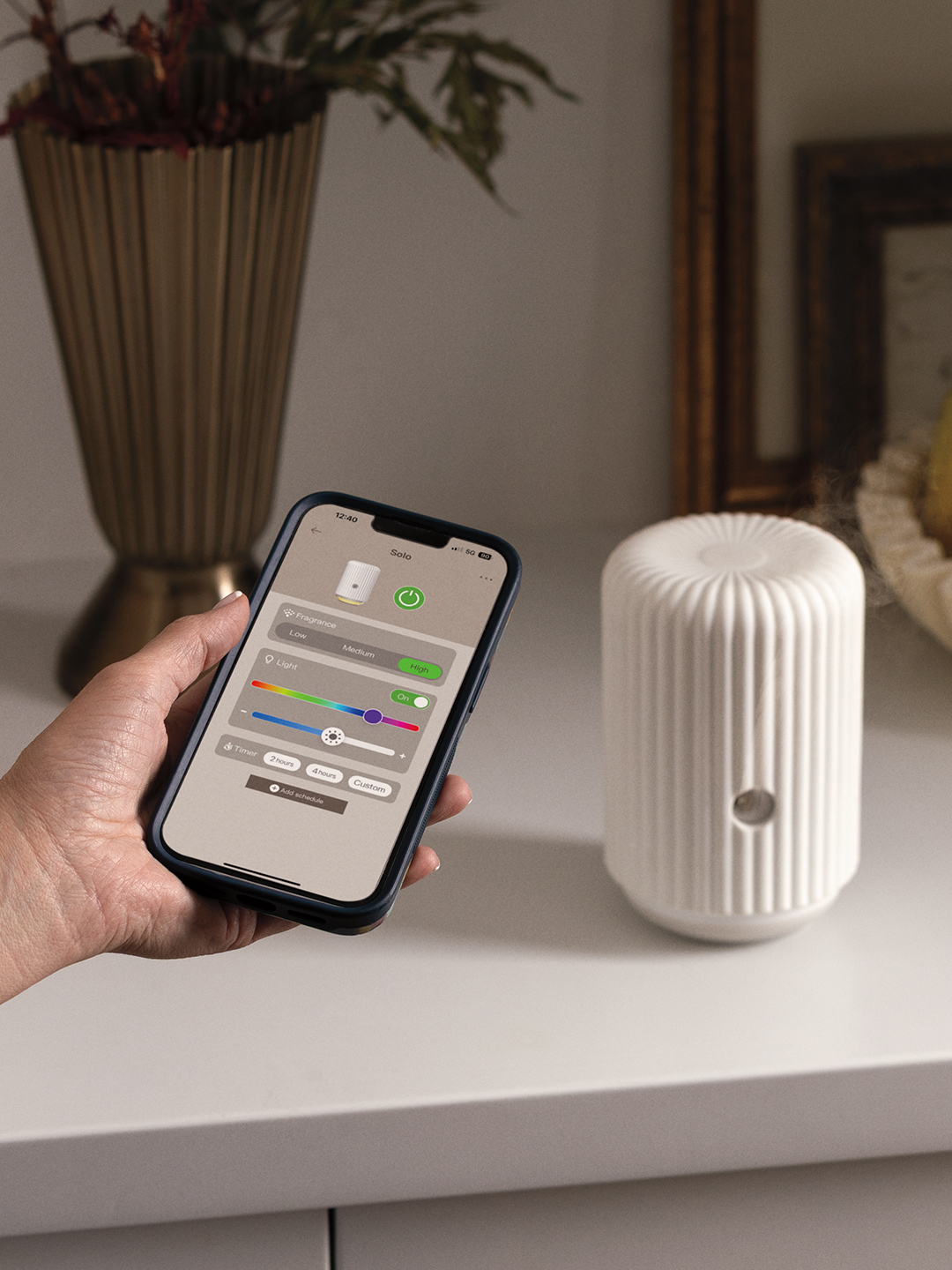 A hand holds a phone showing the Scentsy Solo app controlling fragrance, light, and timer settings, next to the white Scentsy Fragrance Cartridge Diffuser - Solo on a console table.