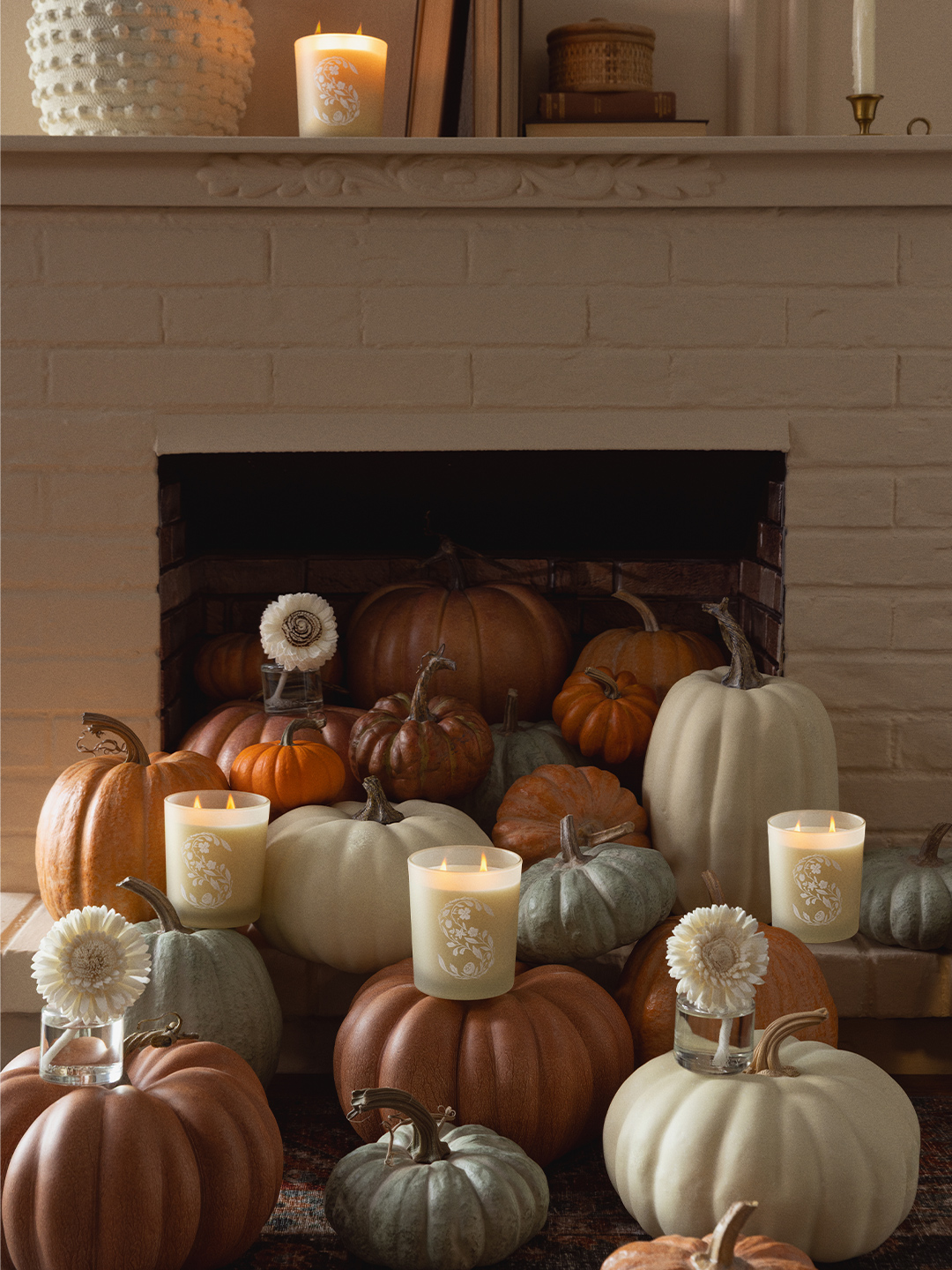 Three lit Scentsy candles in frosted glass jars with a white floral pattern, styled on a fireplace hearth and mantel surrounded by an assortment of orange, white, and sage green decorative pumpkins, dried flower stems in bud vases, and autumn accents.