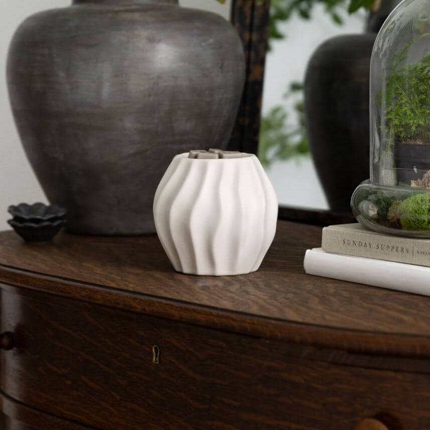 Cascade Warmer in matte white ceramic with soft sculpted curves, styled on a wooden console table beside greenery and decorative accents in a warm, neutral home setting.