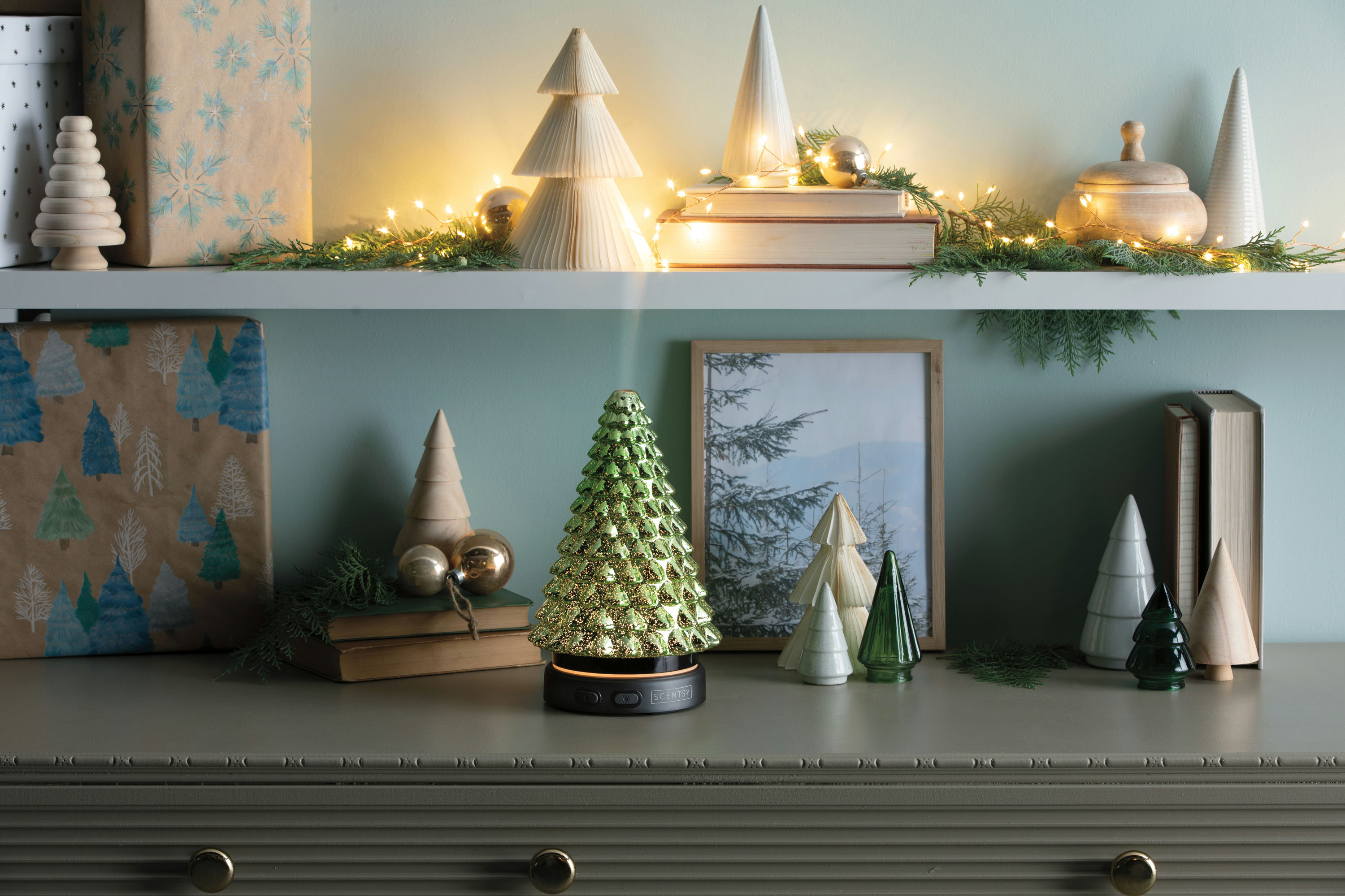 A green mercury-glass Noble & Bright diffuser lit and glowing on a mantel styled with wooden and ceramic holiday trees, wrapped gifts, pine garland, and warm string lights.