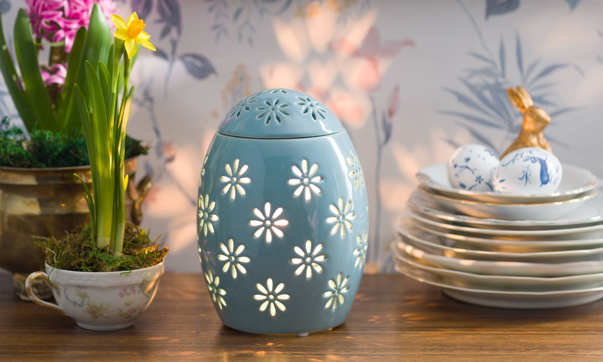 An Easter Garden – Blue Warmer is illuminated on a cabinet beside potted flowers, decorative dishes and Easter Eggs.