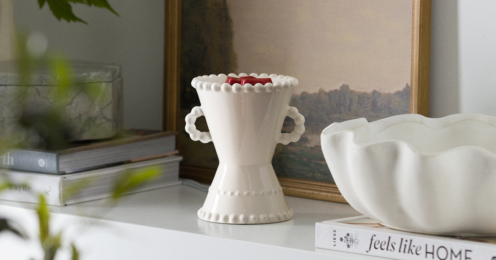 Belaire Warmer in glossy white ceramic with scalloped edges and looped side handles, styled on a shelf with stacked books and framed artwork in a soft, neutral home setting.