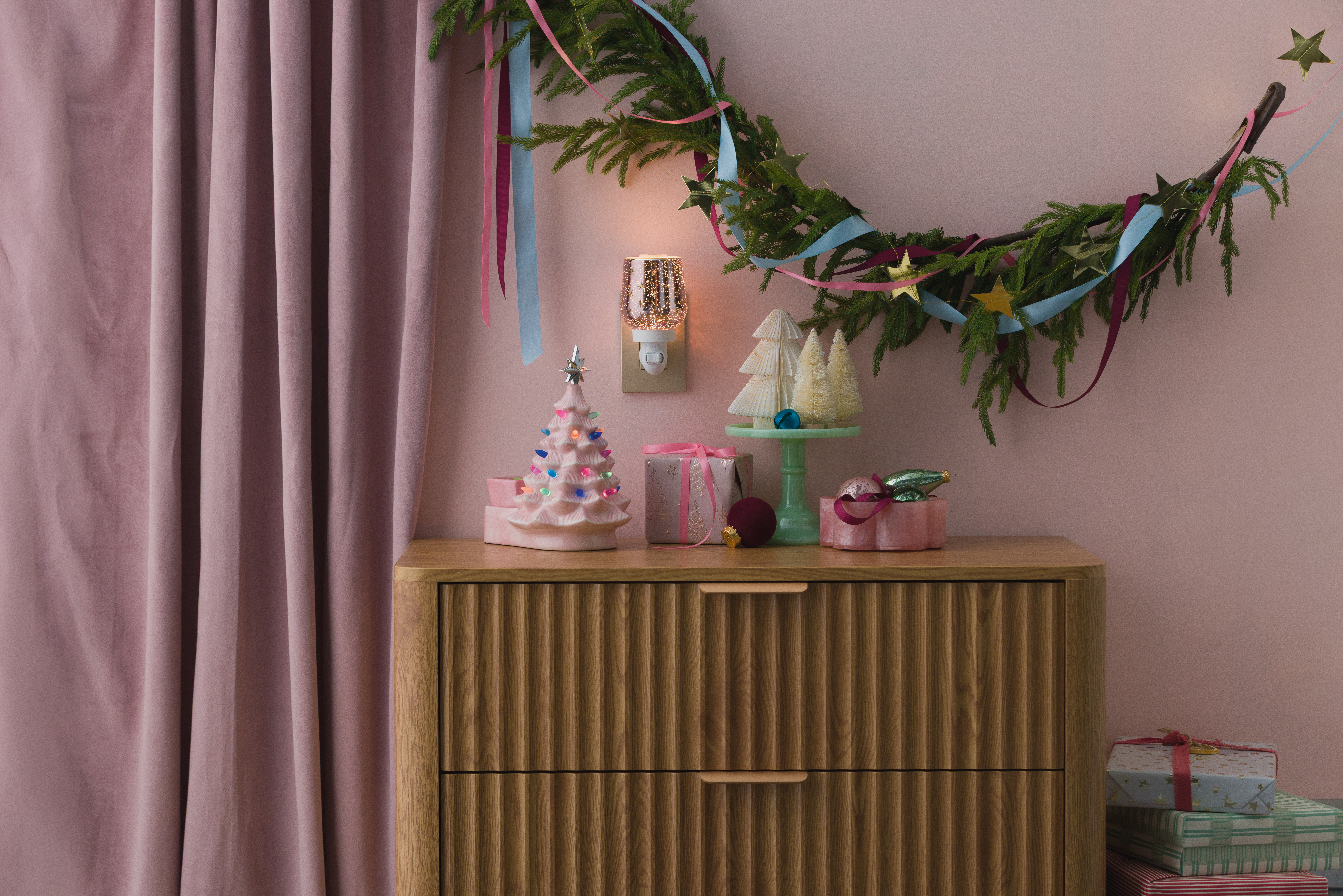 Se muestran un Calentador Merry Little Christmas – Rosa y un Mini Calentador Frosted Flurry en una mesa con decoraciones navideñas en un entorno color rosa.