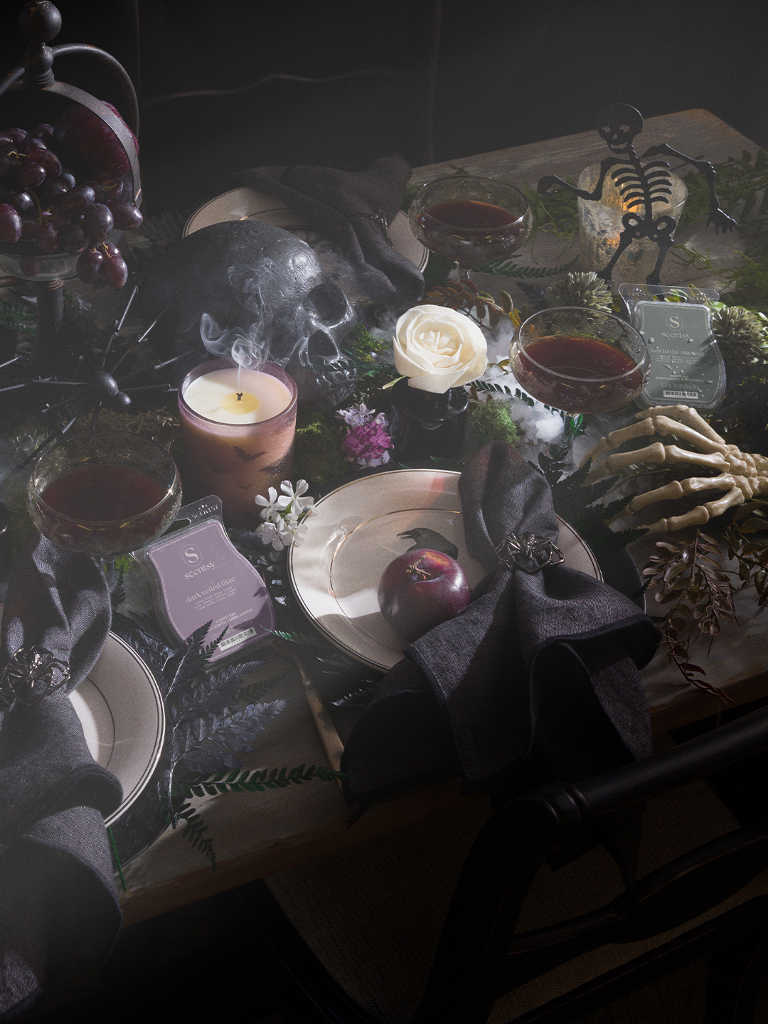 A moody Halloween tabletop styled with a lit Black Luna Night candle in a smoky purple glass jar, a skull, wine-filled glasses, a skeleton hand, a white rose, dried florals, and Scentsy wax bars including Dark Veiled Lilac and Lost in the Amazon, set against dark place settings with black napkins.
