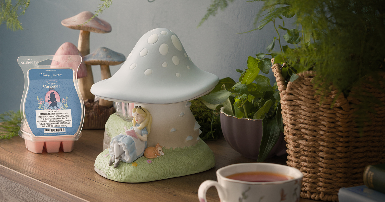 Un Calentador Scentsy – Disney Alice in Wonderland: Down the Rabbit Hole está junto a una Barra Scentsy – Disney Alice in Wonderland: Curiouser and Curiouser abierta en una mesa con plantas, setas decorativas y una taza de té.