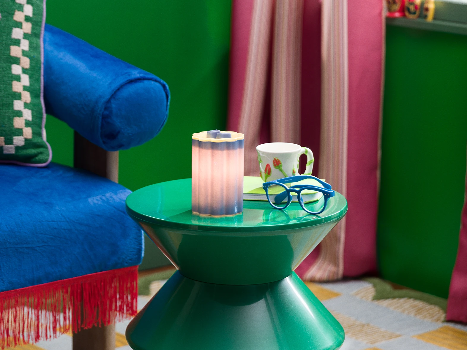 A Petal – Purple Warmer is displayed on a white tray with brightly coloured vases, tulips and a green cabinet in the background. 