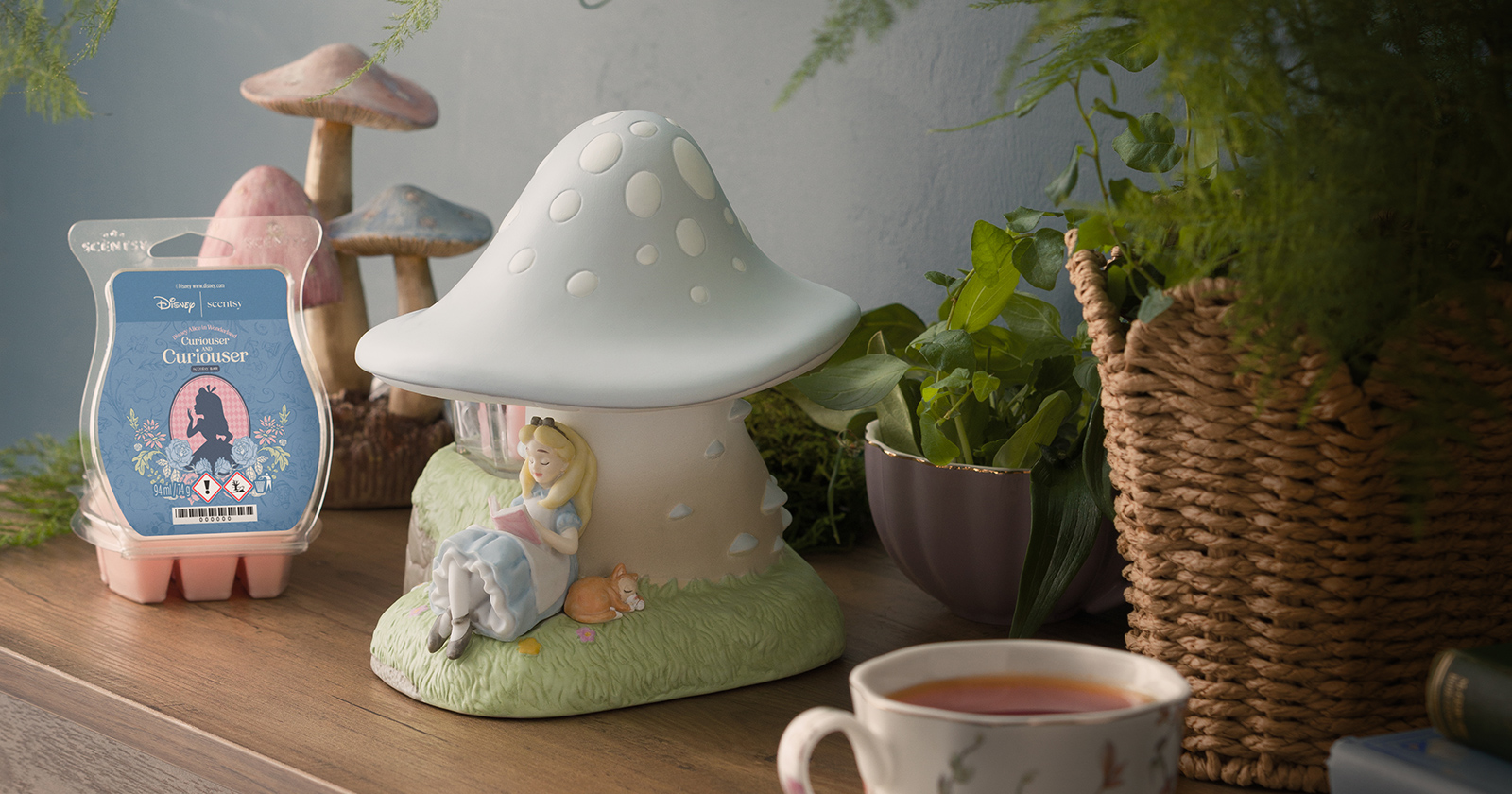 A Disney Alice in Wonderland: Down the Rabbit Hole – Scentsy Warmer sits next to an open Disney Alice in Wonderland: Curiouser and Curiouser – Scentsy Bar on a table with plants, decorative mushrooms and a cup of tea.