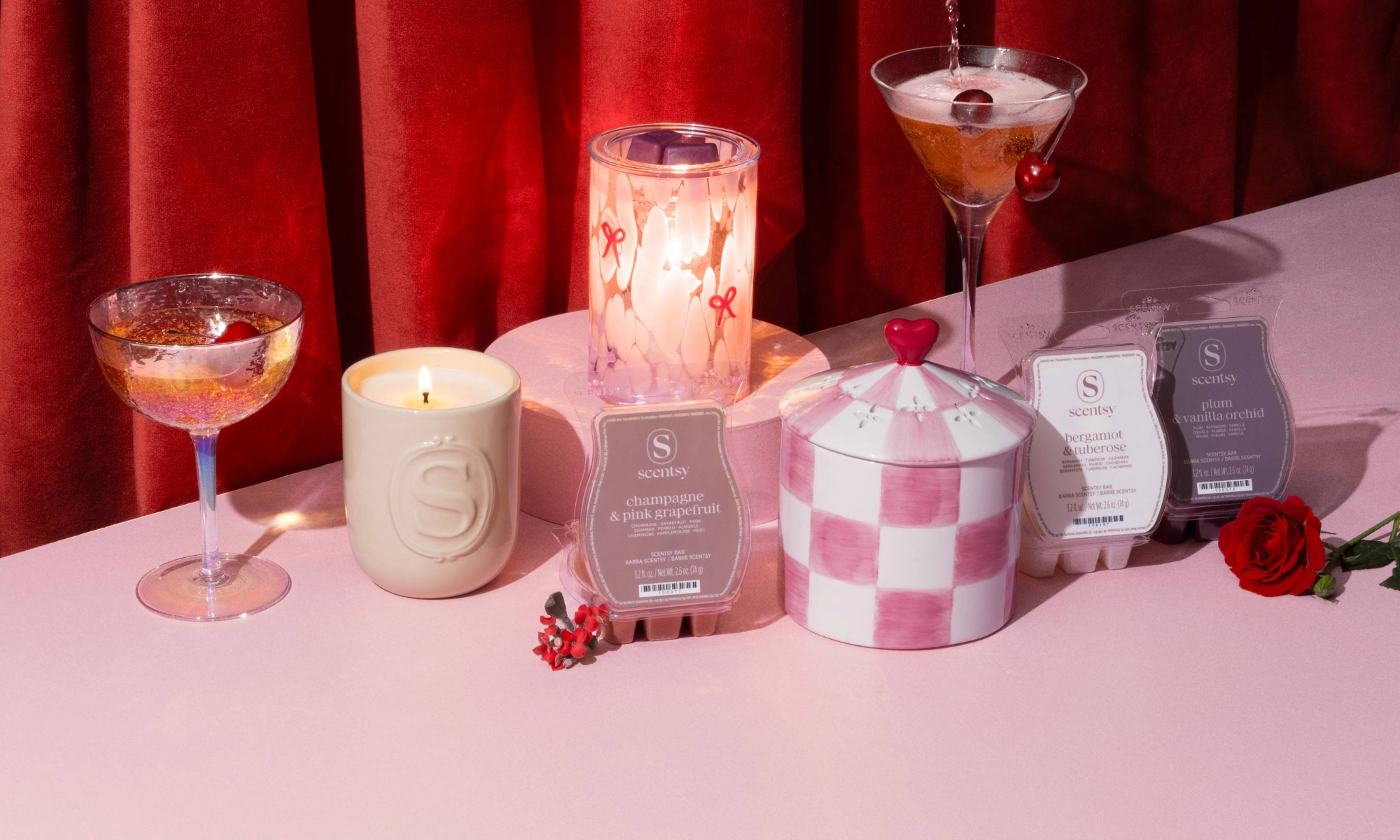 Valentine’s Day Collection featuring the Adorned Affair Warmer, First Blush Warmer, Valentine’s Day 3-Bar Bundle, and Petals & Poems Scented Candle styled with pink cocktails in front of red velvet curtains.