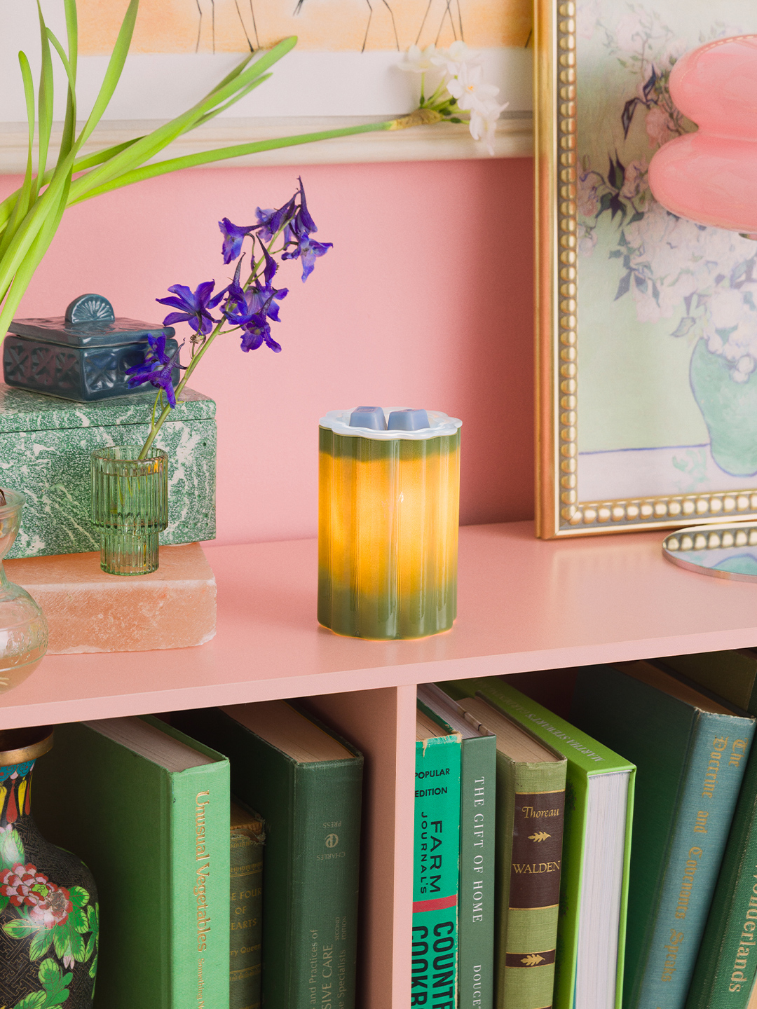 Bloomie-Green glass warmer lit to reveal a soft yellow-to-green ombré glow, styled on a pink shelf beside books, flowers, and framed art.