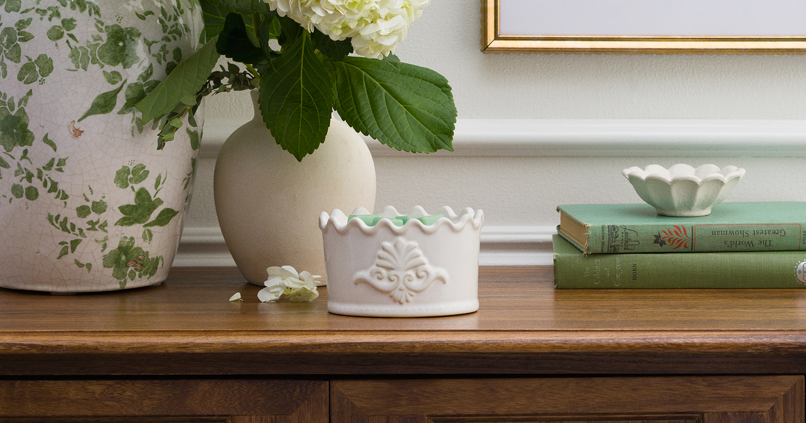 Classic Crest Warmer in white ceramic with embossed detailing, styled on a wooden surface beside greenery and framed artwork in a soft, neutral home setting.
