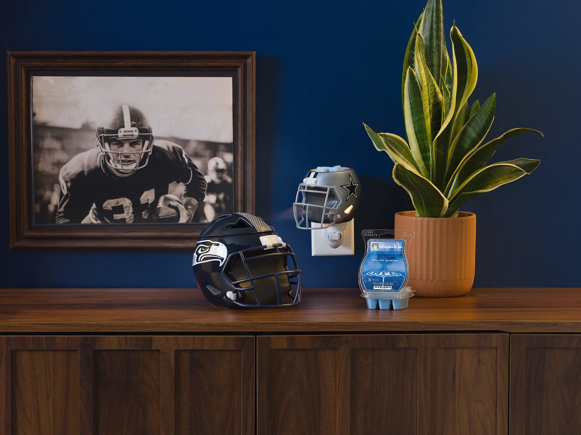 An NFL: Seattle Seahawks Helmet – Scentsy Warmer and an NFL: Dallas Cowboys Helmet – Scentsy Mini Warmer are displayed with an open NFL: 4th Quarter – Scentsy Bar near a plant and framed photo. 