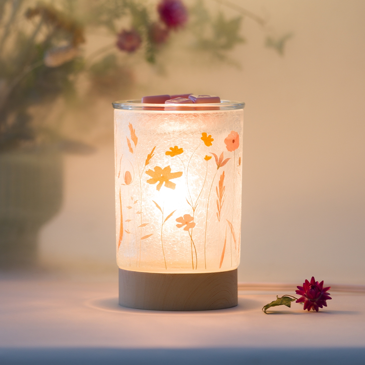 Calentador Pick of the Bunch de Scentsy encendido, con diseño de flores prensadas en tonos amarillo y coral sobre vidrio esmerilado con base de madera clara, se muestra con cera derritiéndose arriba y una flor seca rosa a un lado.