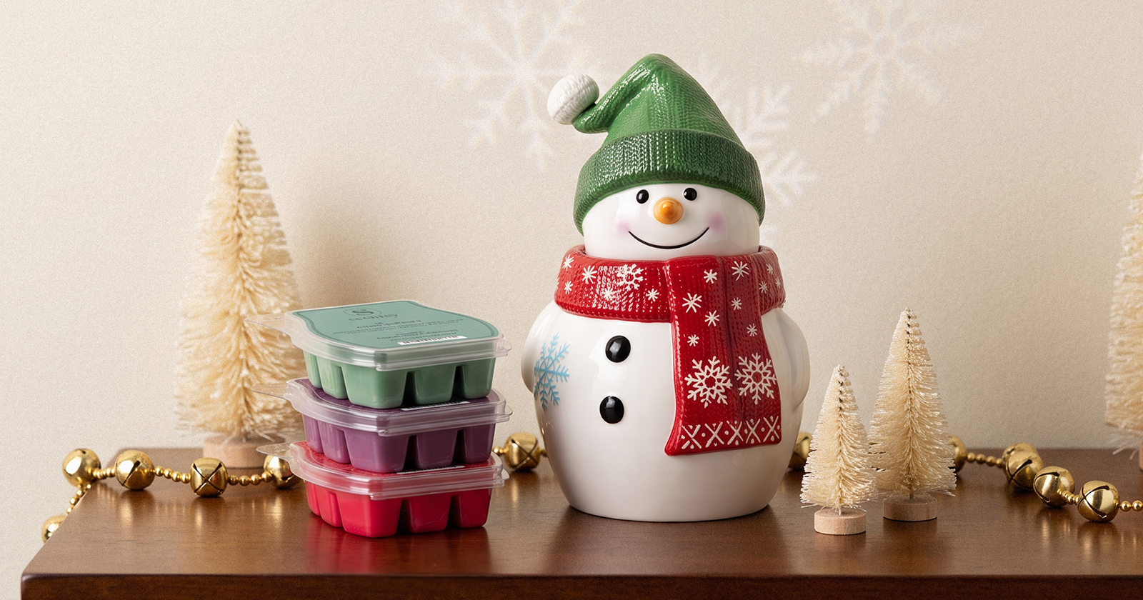 A snowman-shaped Scentsy Warmer wearing a green knit hat and red snowflake scarf is displayed with a stack of Scentsy Bars, bottlebrush trees, and gold jingle bells.