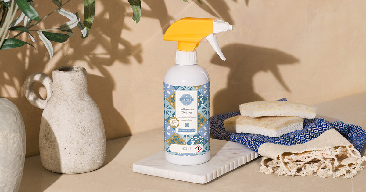 Scentsy bathroom cleaner spray bottle with yellow trigger top and blue Mediterranean tile pattern label on white marble tray, styled with ceramic vases, towels, and olive branches