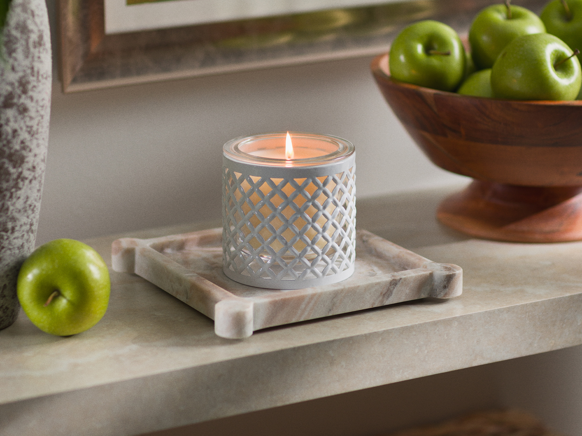 A lit Scentsy Candle Insert sits in a white trellis-pattern Scentsy Candle Holder on a marble tray, with green apples nearby.