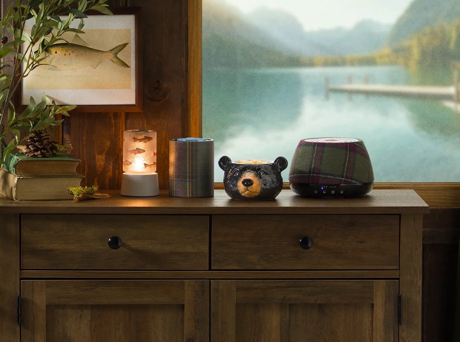 Cozy Cabin Edit Warmers on display in a rustic cabin setting with evergreen and pinecones, a fish painting and a window to the lake outside.