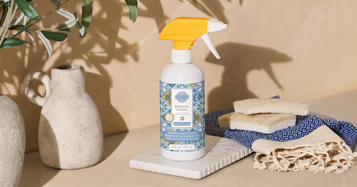 Scentsy bathroom cleaner spray bottle with yellow trigger top and blue Mediterranean tile pattern label on white marble tray, styled with ceramic vases, towels, and olive branches