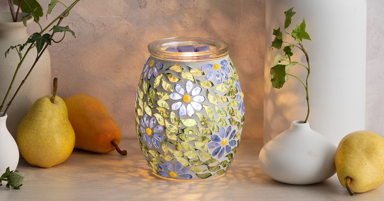 Scentsy Warmer with purple and white floral mosaic pattern and yellow centers, illuminated from within, displayed on wood surface with pears and white ceramic vases