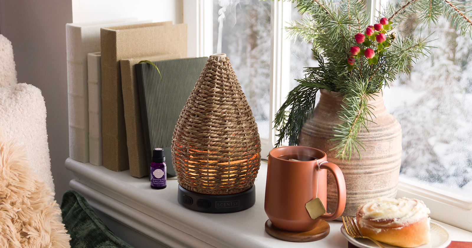 Scentsy Wonder woven rattan teardrop essential oil diffuser with warm amber glow on white windowsill, displayed with terracotta mug, pine branch arrangement, books, and frosted cinnamon roll