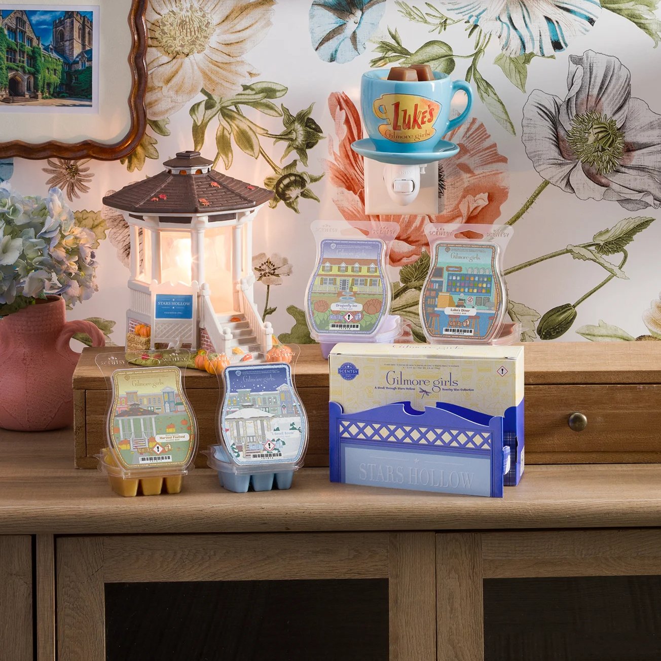 The Gilmore Girls: Welcome to Stars Hollow – Scentsy Warmer and Scentsy Bars from the A Stroll Through Stars Hollow – Scentsy Wax Collection are arranged on a table. Behind it, the Luke’s Coffee Mug – Scentsy Mini Warmer is plugged into a wall with floral wallpaper.