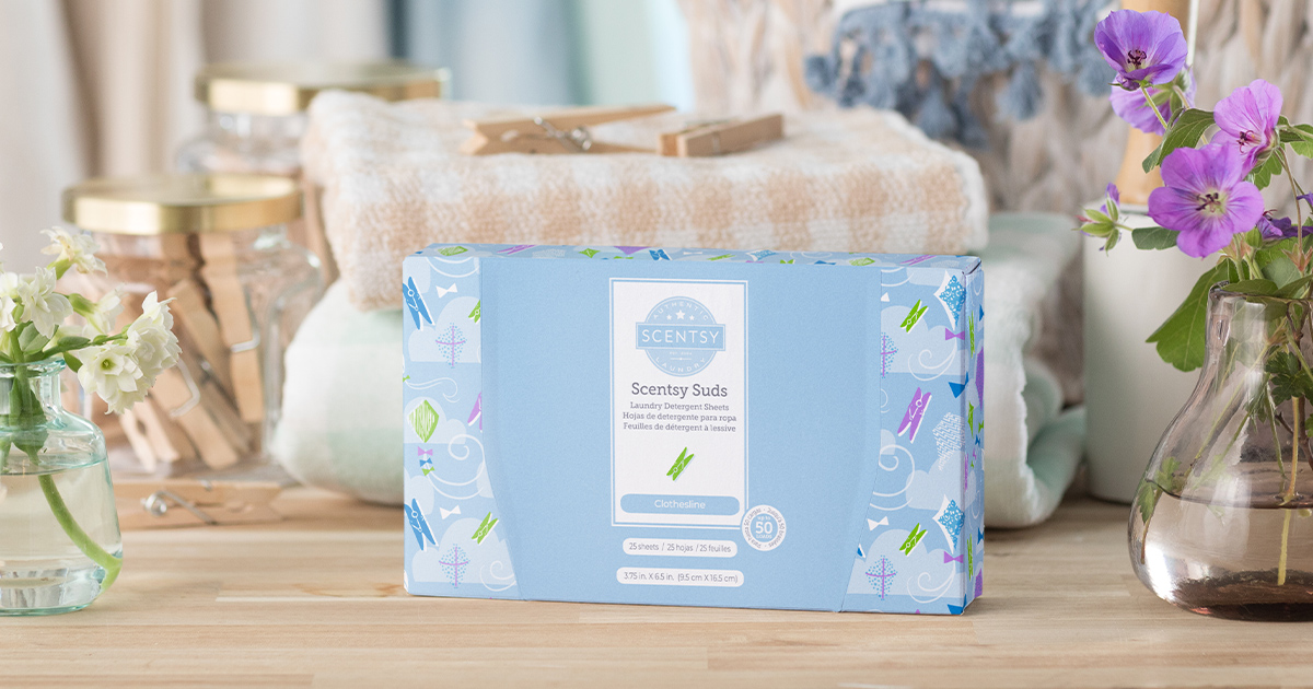Scentsy Suds Clothesline laundry detergent sheets in light blue packaging, displayed on wood surface with folded towels, glass jars, and purple flowers