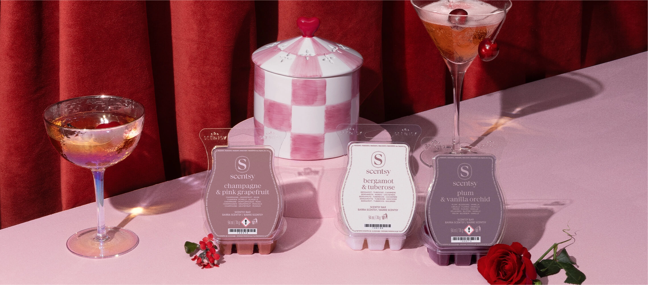 Valentine’s Day Collection featuring the Adorned Affair Warmer, First Blush Warmer, Valentine’s Day 3-Bar Bundle, and Petals & Poems Scented Candle styled with pink cocktails in front of red velvet curtains.