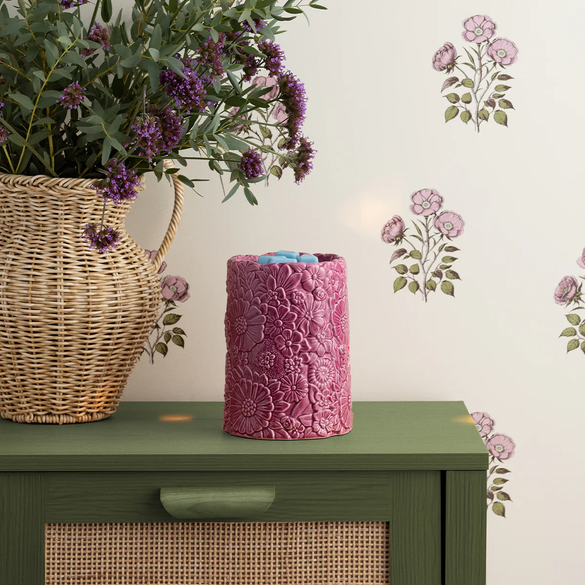 The Dusky Petals Scentsy Warmer is shown on a side table next to flowers in a wicker vase.