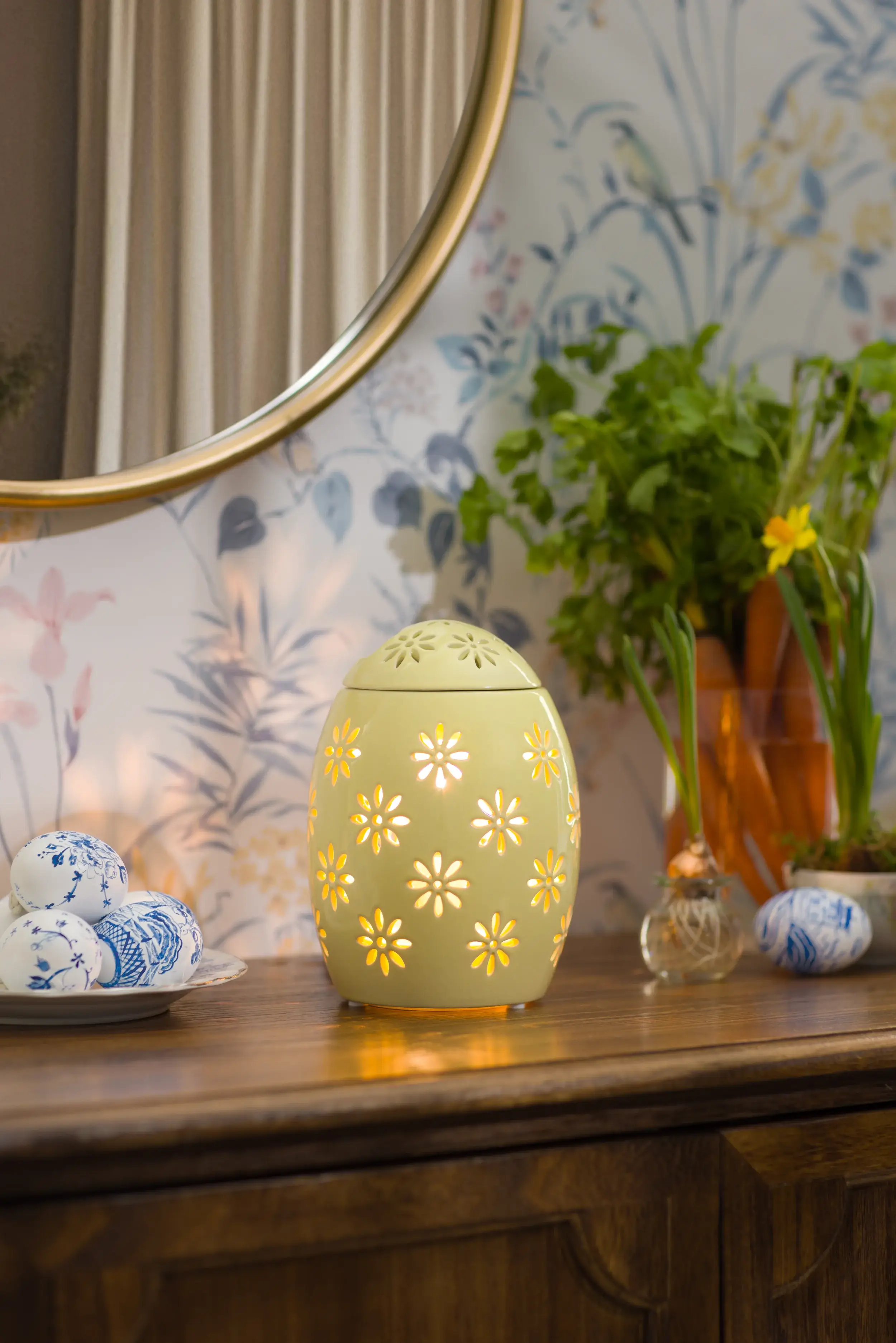 Un Réchaud Easter Garden – Yellow est allumé sur un meuble à côté de plantes d’intérieur et d’œufs de Pâques.