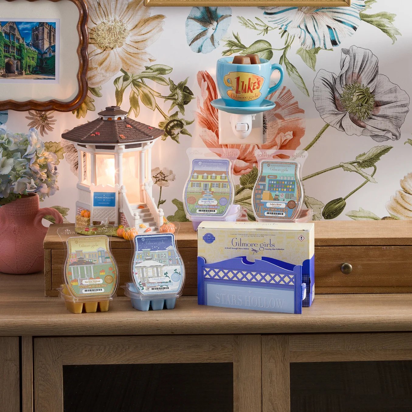 The Gilmore Girls: Welcome to Stars Hollow – Scentsy Warmer and Scentsy Bars from the A Stroll Through Stars Hollow – Scentsy Wax Collection are arranged on a table. Behind it, the Luke’s Coffee Mug – Scentsy Mini Warmer is plugged into a wall with floral wallpaper.