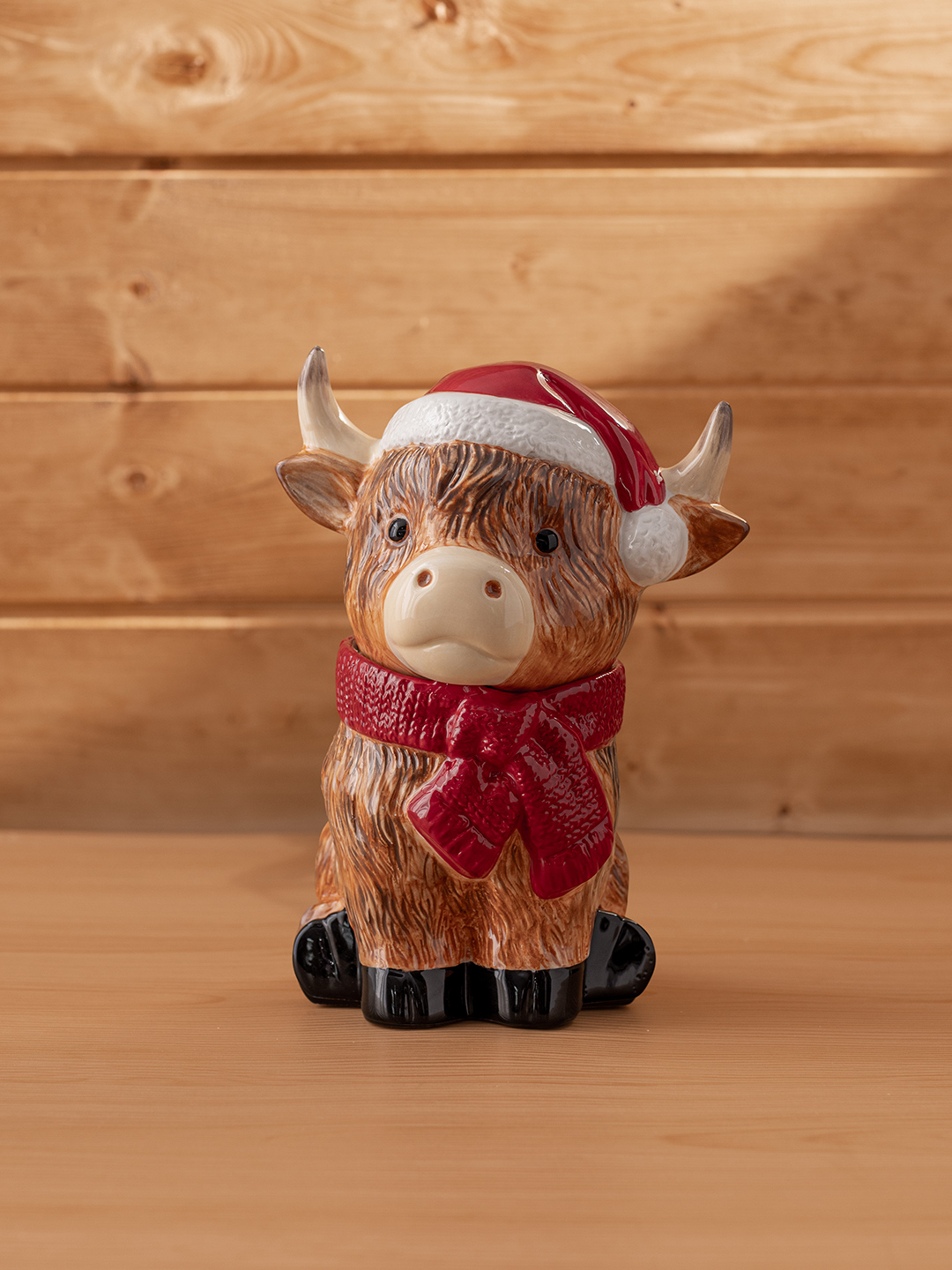 A Jolly Ol’ Saint Moo Warmer wearing a Santa hat and red scarf is displayed on a wooden surface.