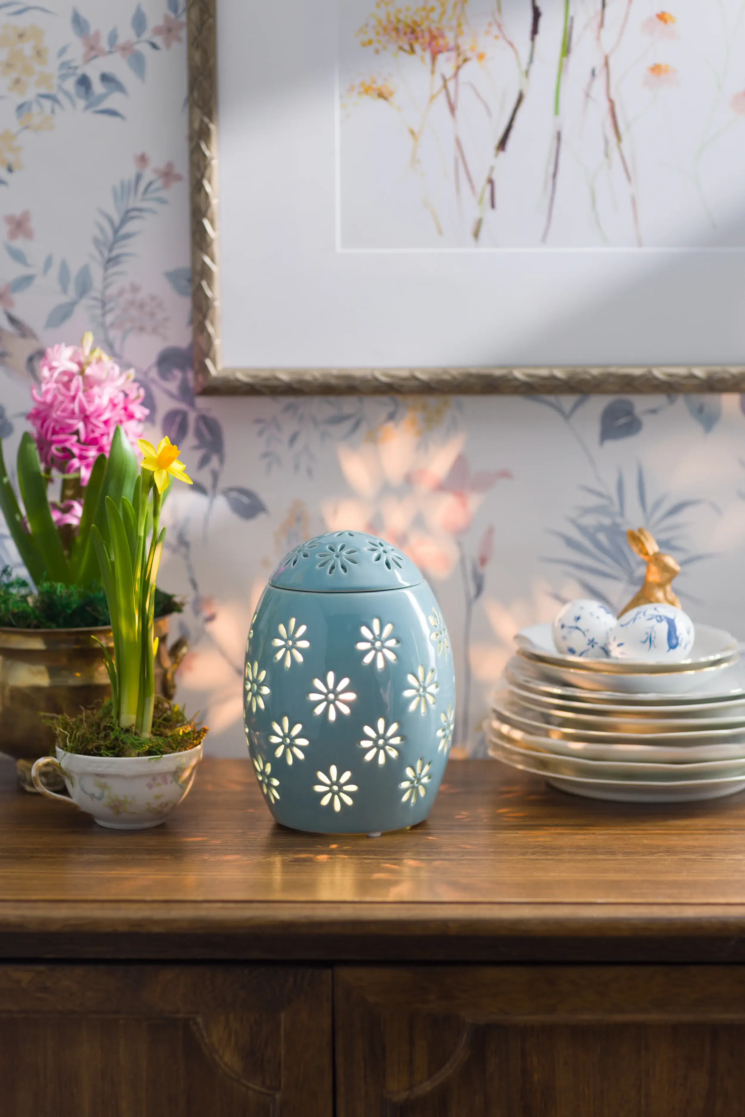 Un Réchaud Easter Garden – Blue est allumé sur un meuble à côté de fleurs en pot, de plats décoratifs et d’œufs de Pâques.