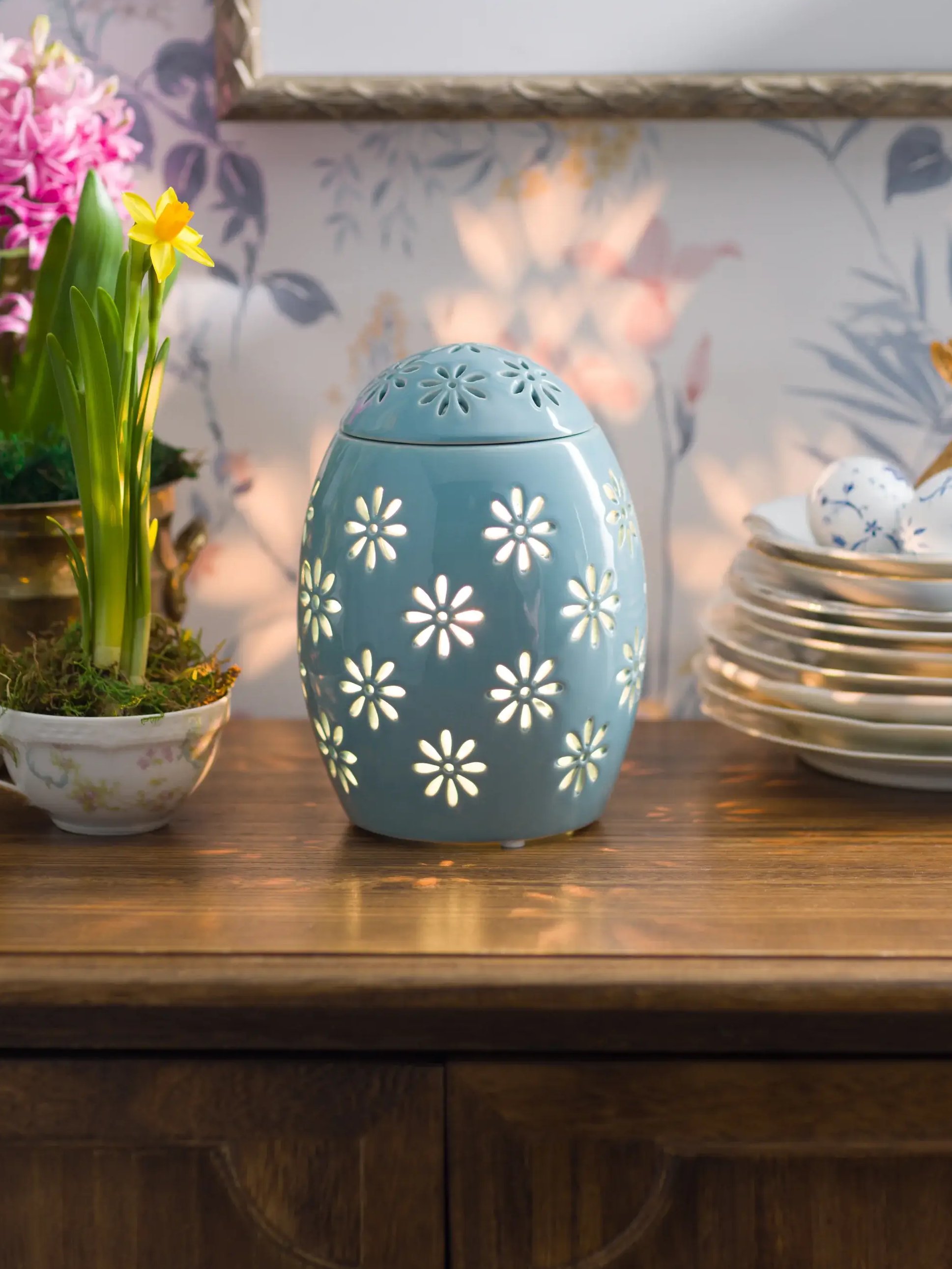 An Easter Garden – Blue Warmer is illuminated on a cabinet beside potted flowers, decorative dishes and Easter Eggs.