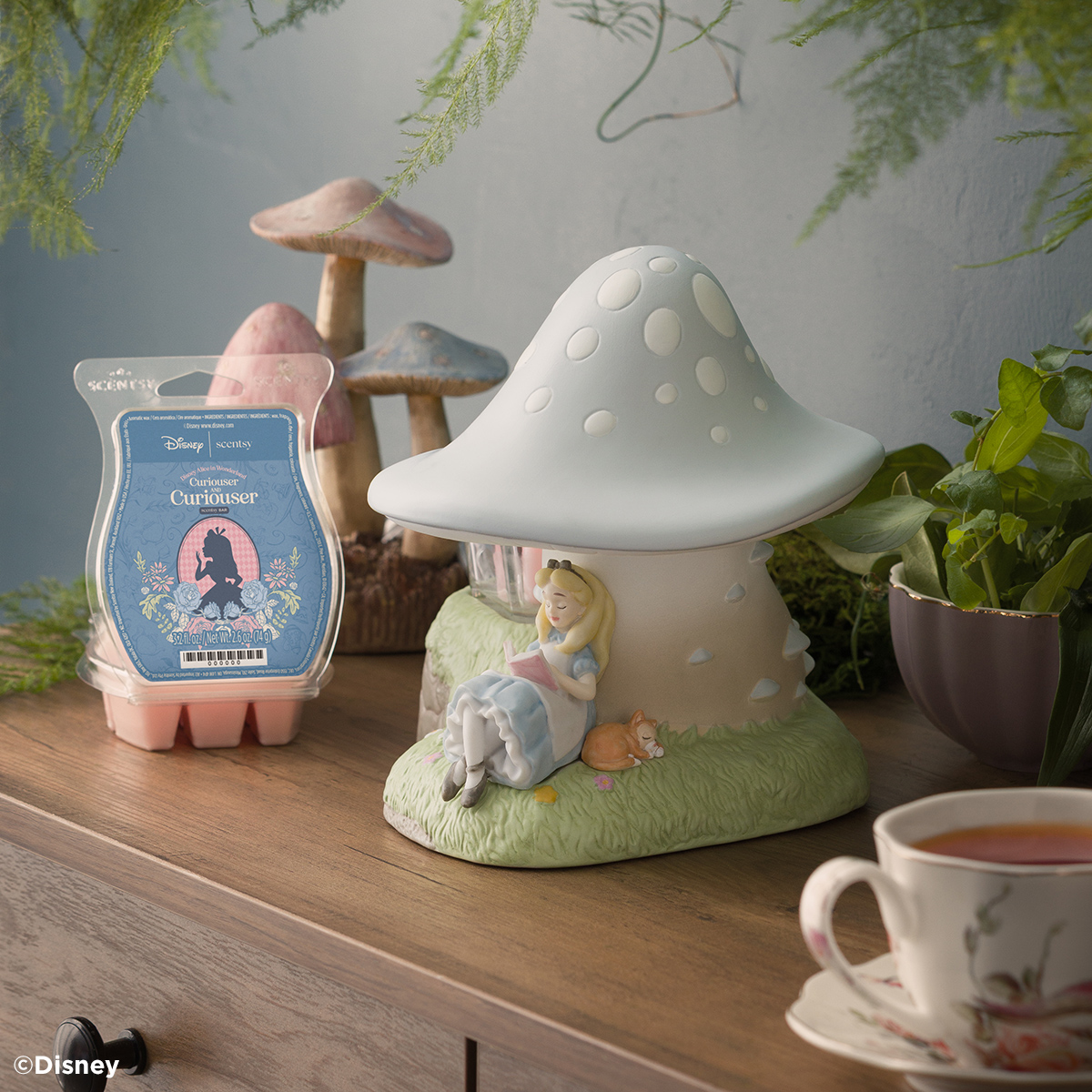 A Disney Alice in Wonderland: Down the Rabbit Hole – Scentsy Warmer sits next to an open Disney Alice in Wonderland: Curiouser and Curiouser – Scentsy Bar on a table with plants, decorative mushrooms and a cup of tea.