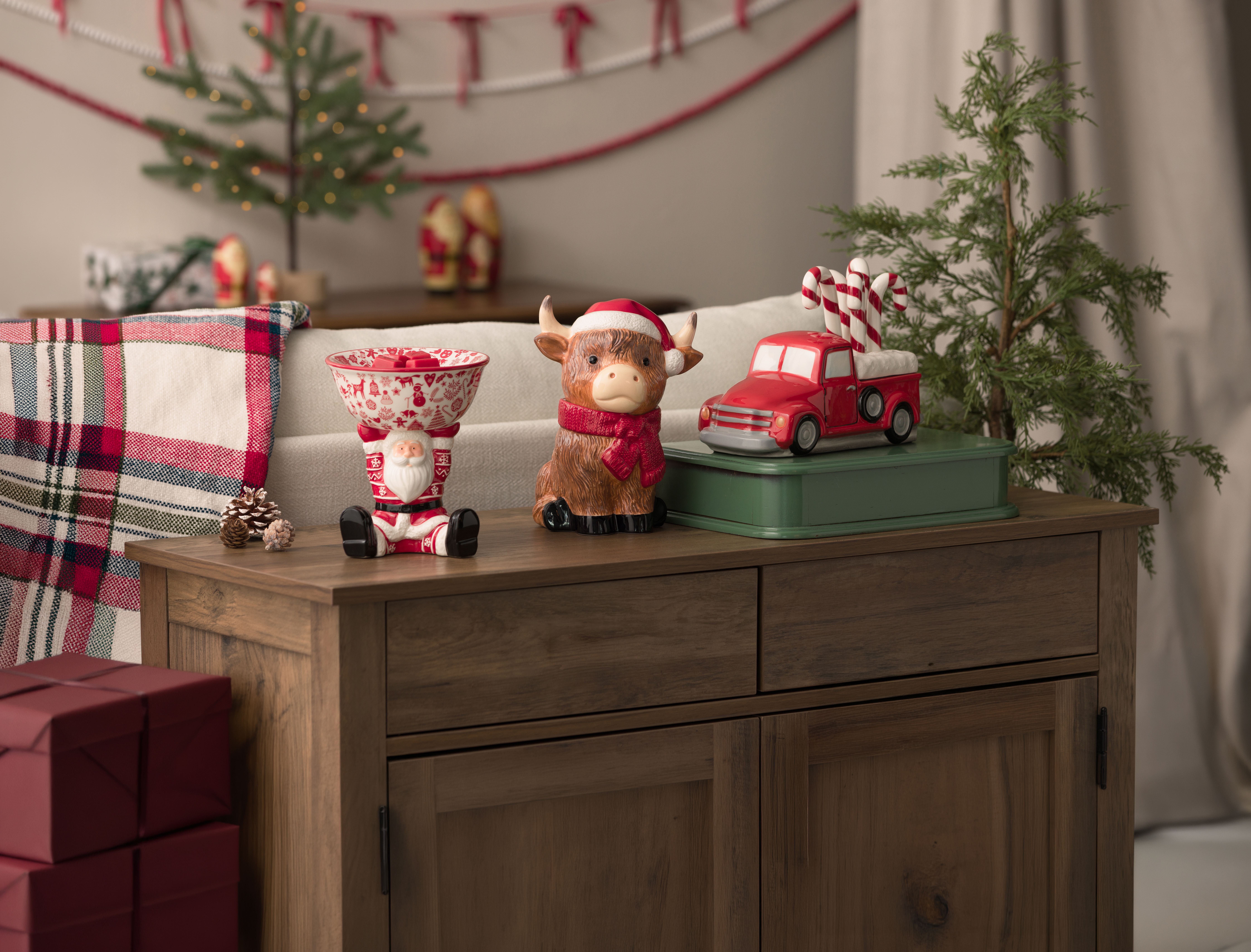 Eine Elektrische Duftlampe Christmas Keeper, eine Elektrische Duftlampe Jolly Ol’ Saint Moo und eine Elektrische Duftlampe Red Retro Truck mit einem Zuckerstangendeckel stehen in einem weihnachtlich geschmückten Wohnzimmer.