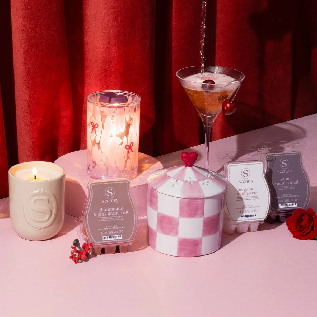 Valentine’s Day Collection products displayed on a pink table among flowers and glasses being filled with a sparkling beverage, all in front of a red curtain.