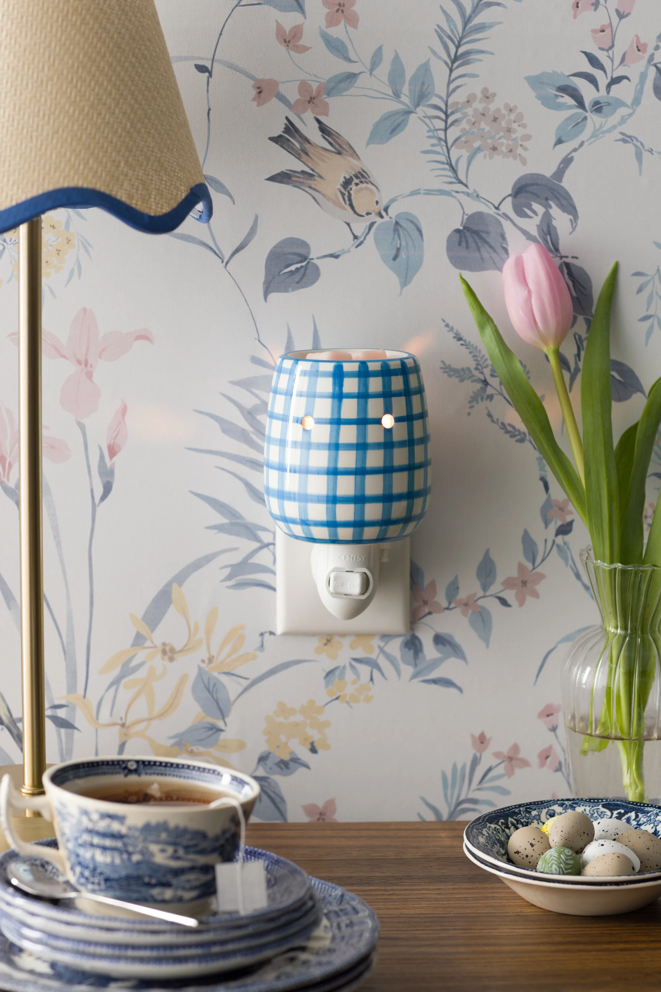 A Cottage Gingham – Blue Mini Warmer is plugged into a floral-patterned wall beside a vase of tulips.