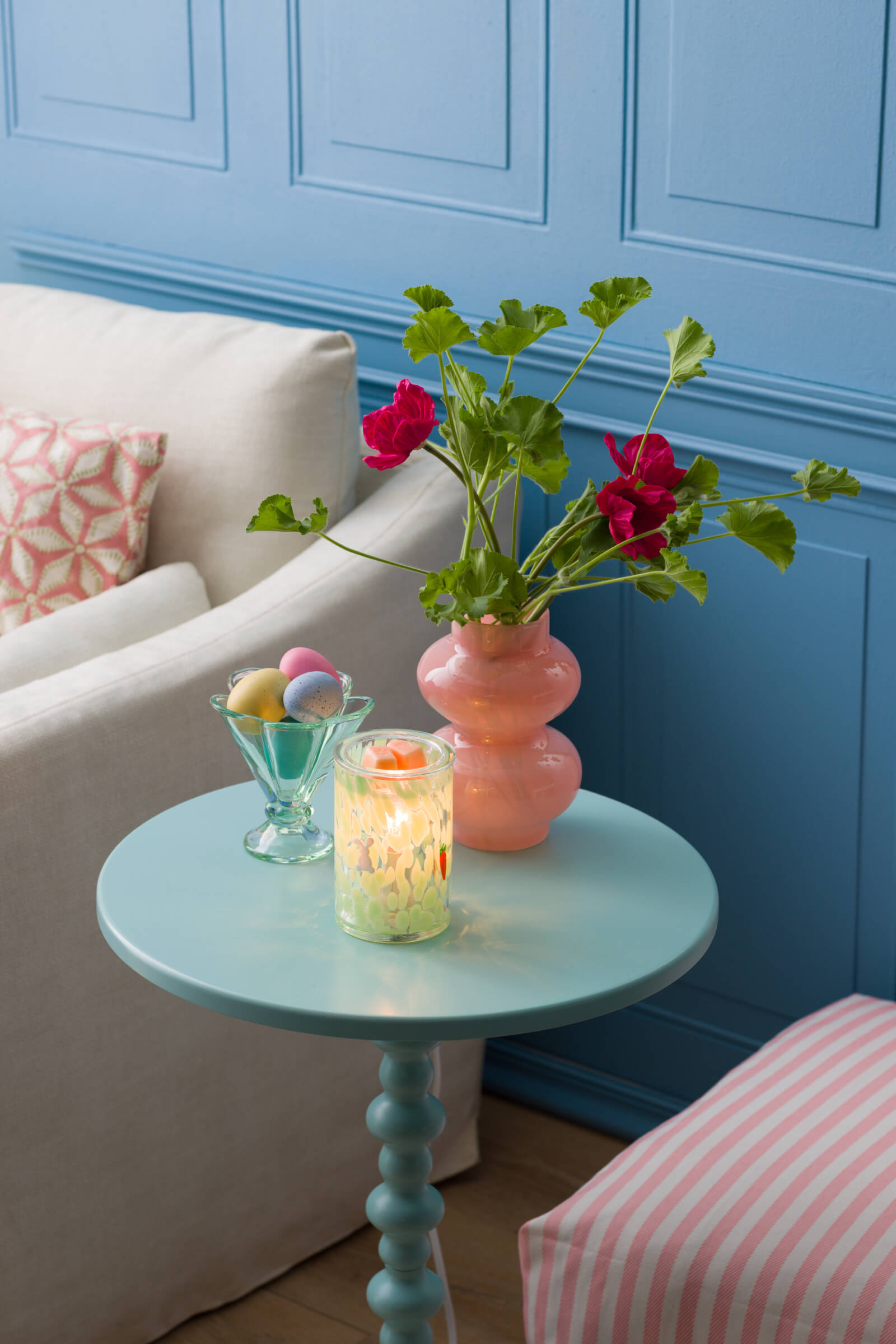 A lit Verdant Escape Warmer sits on a pastel end table beside Easter eggs and a vase of flowers.